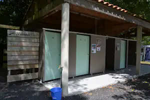 Sanitärgebäude am Campingplatz mit überdachtem Eingang, grünen Türen, Waschbereich und Schildern an der Wand.