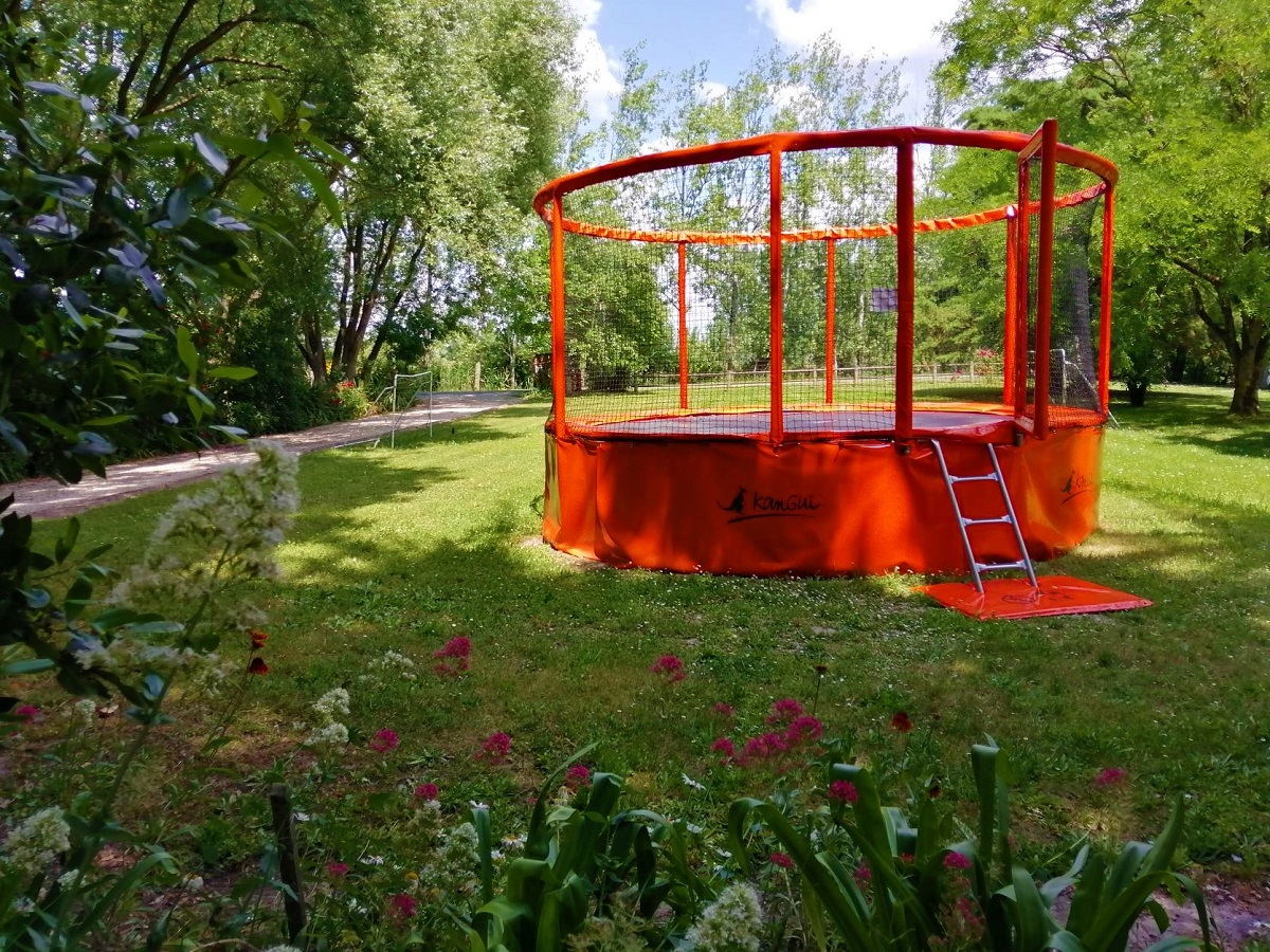 Ein Trampolin mit Schutznetz im grünen Campinggarten, umgeben von Rasen, Blumenbeeten und Bäumen.