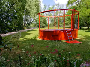 Ein Trampolin mit Schutznetz im grünen Campinggarten, umgeben von Rasen, Blumenbeeten und Bäumen.