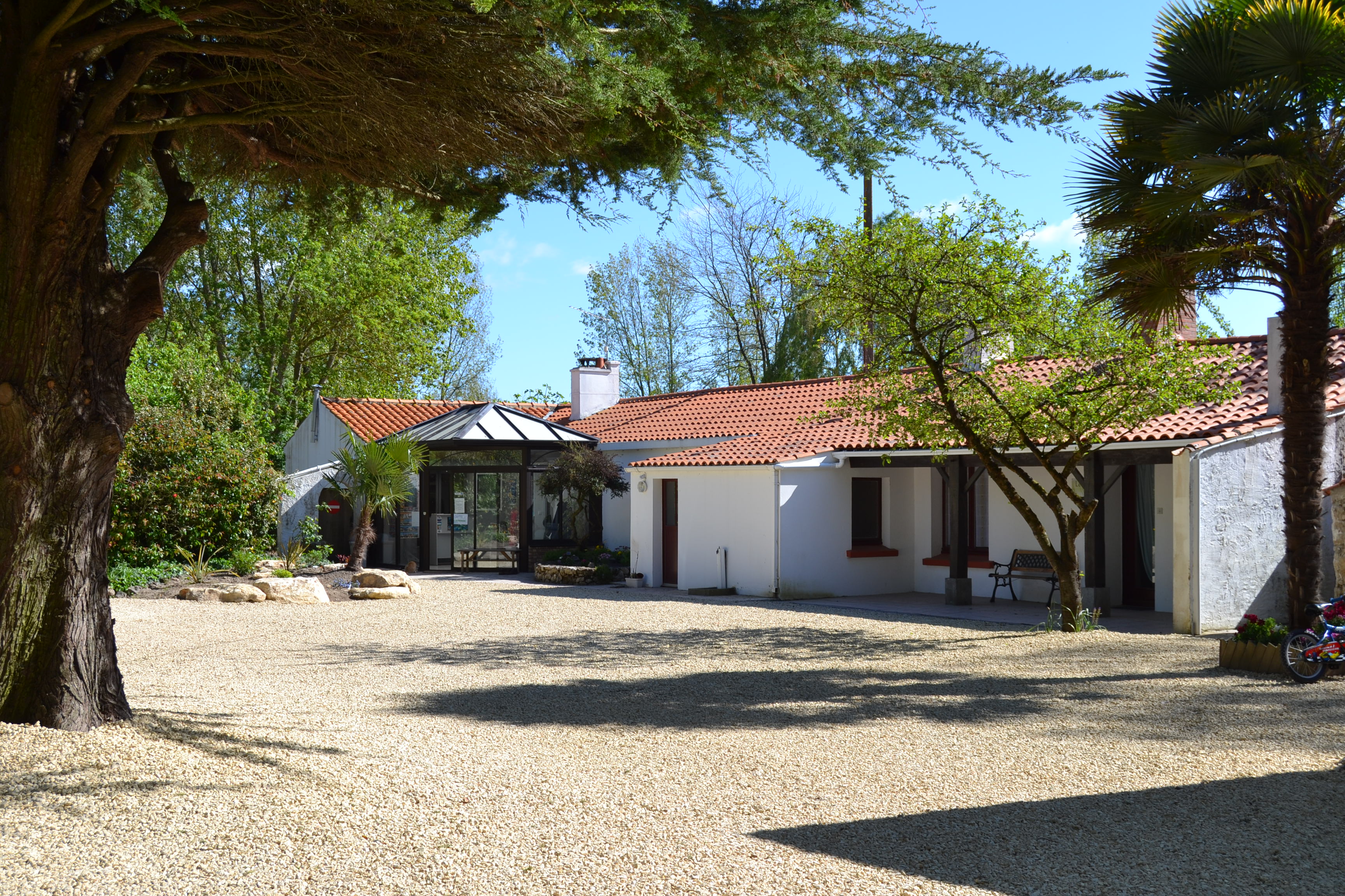 Weißes Ferienhaus mit Terrasse, Ziegel­dach und Schornstein; Kiesweg und Bäume im Vordergrund.