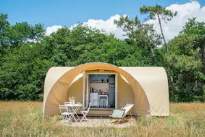 Beiges Zelt auf einem Campingplatz im Gras, mit zwei Stühlen und offenem Eingang vor dichtem Grün und Bäumen.