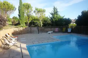 Privater Pool mit Liegestühlen und weißen Gartenstühlen auf gepflasterter Terrasse, umgeben von Bäumen und Hecken.