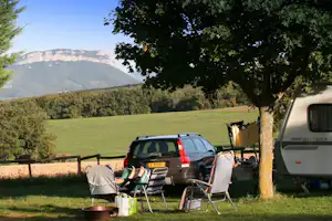 Campingplatz mit Wohnwagen und Auto, Campingstühle am Tisch unter einem Baum, Wiese und Berge im Hintergrund.