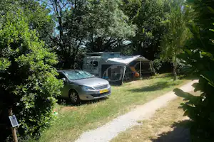 Wohnwagen mit Vorzelt und geparktem Auto auf einem Grasstellplatz zwischen Bäumen und Hecken am Campingplatz.