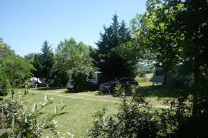 Campingplatz mit Rasenfläche, parkenden Autos und einem Wohnmobil am Rand, umgeben von Bäumen und Hecken.