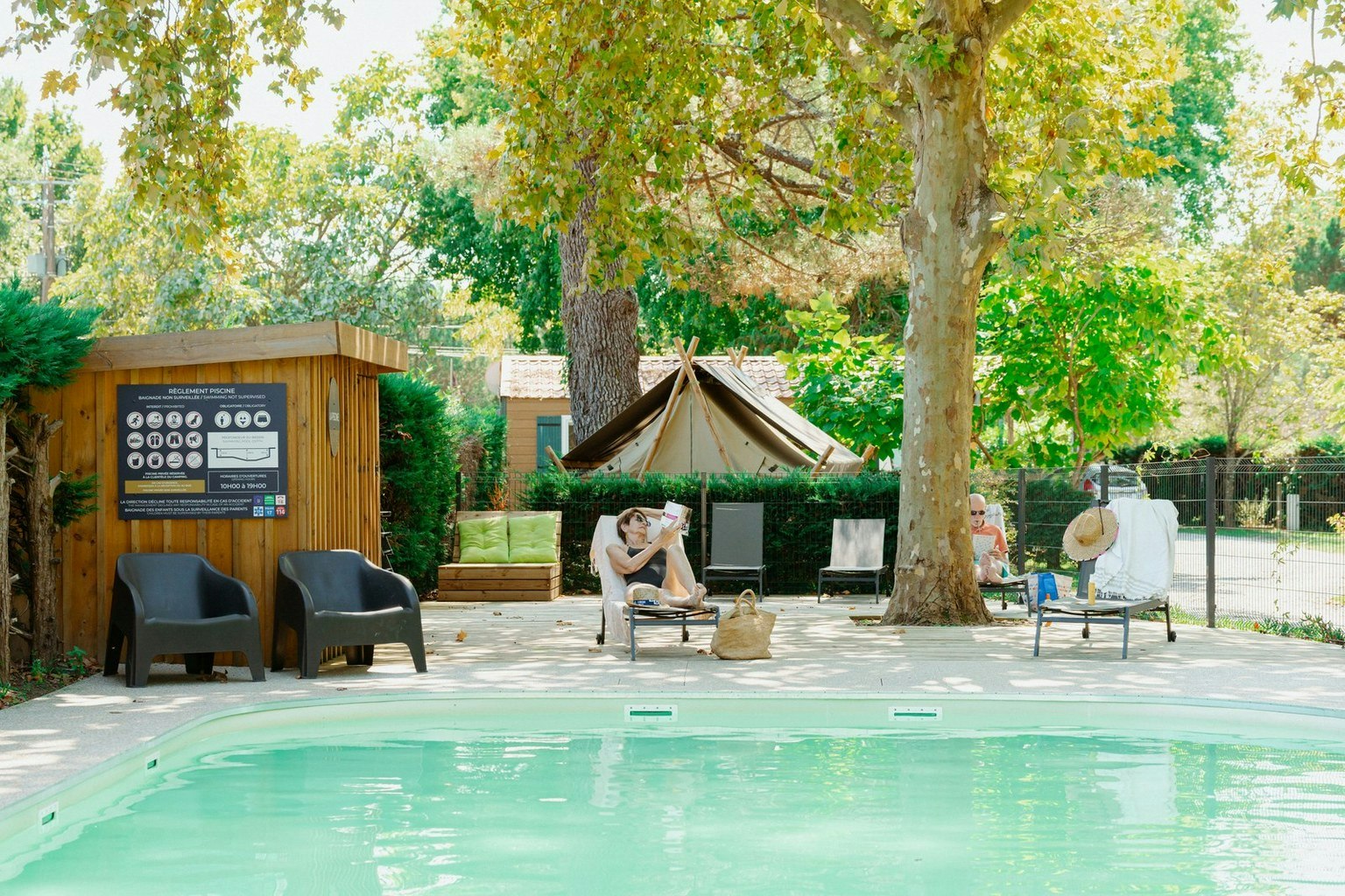 Schwimmbecken mit türkisfarbenem Wasser, daneben Gartenmöbel und ein Zelt im Hintergrund zwischen Bäumen und Hecken.