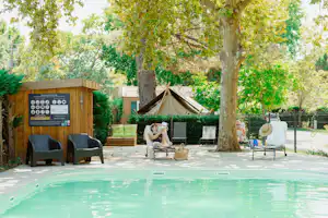 Schwimmbecken mit türkisfarbenem Wasser, daneben Gartenmöbel und ein Zelt im Hintergrund zwischen Bäumen und Hecken.