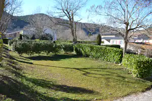 Grüne Campinganlage mit gepflegtem Rasen, Hecken und Bäumen, im Hintergrund Häuser und bewaldete Hügel.