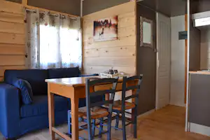 Wohnbereich in einem Holz-Campinghaus: Esstisch mit Stühlen, Sofa, Vorhänge und Wandbilder, daneben eine Tür und Spiegel.