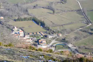Blick auf ein Dorf im Tal mit Häusern, Feldern und einem Fluss, umgeben von bewaldeten Hängen und Straßen.