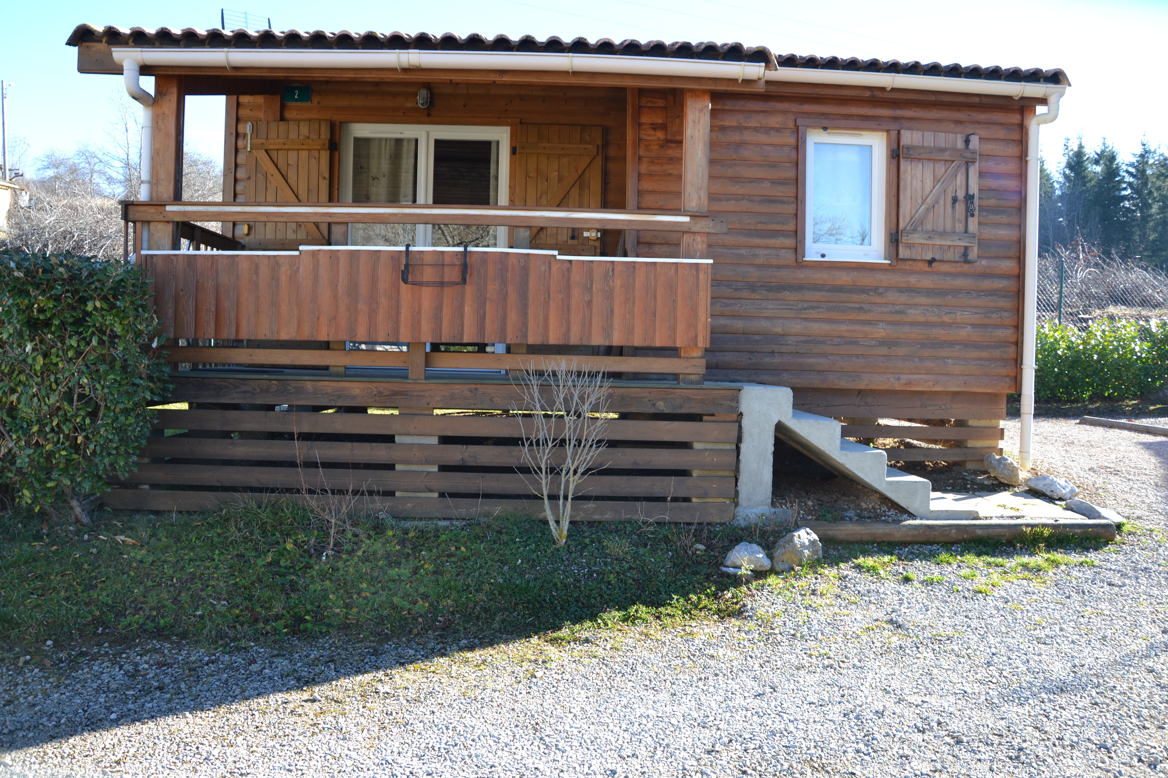 Holzbungalow mit Terrasse und Stufen, Fenster und Eingang, Kiesweg davor und Hecken im Hintergrund.