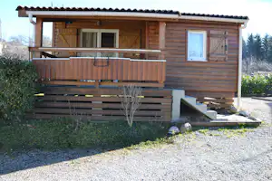 Holzbungalow mit Terrasse und Stufen, Fenster und Eingang, Kiesweg davor und Hecken im Hintergrund.