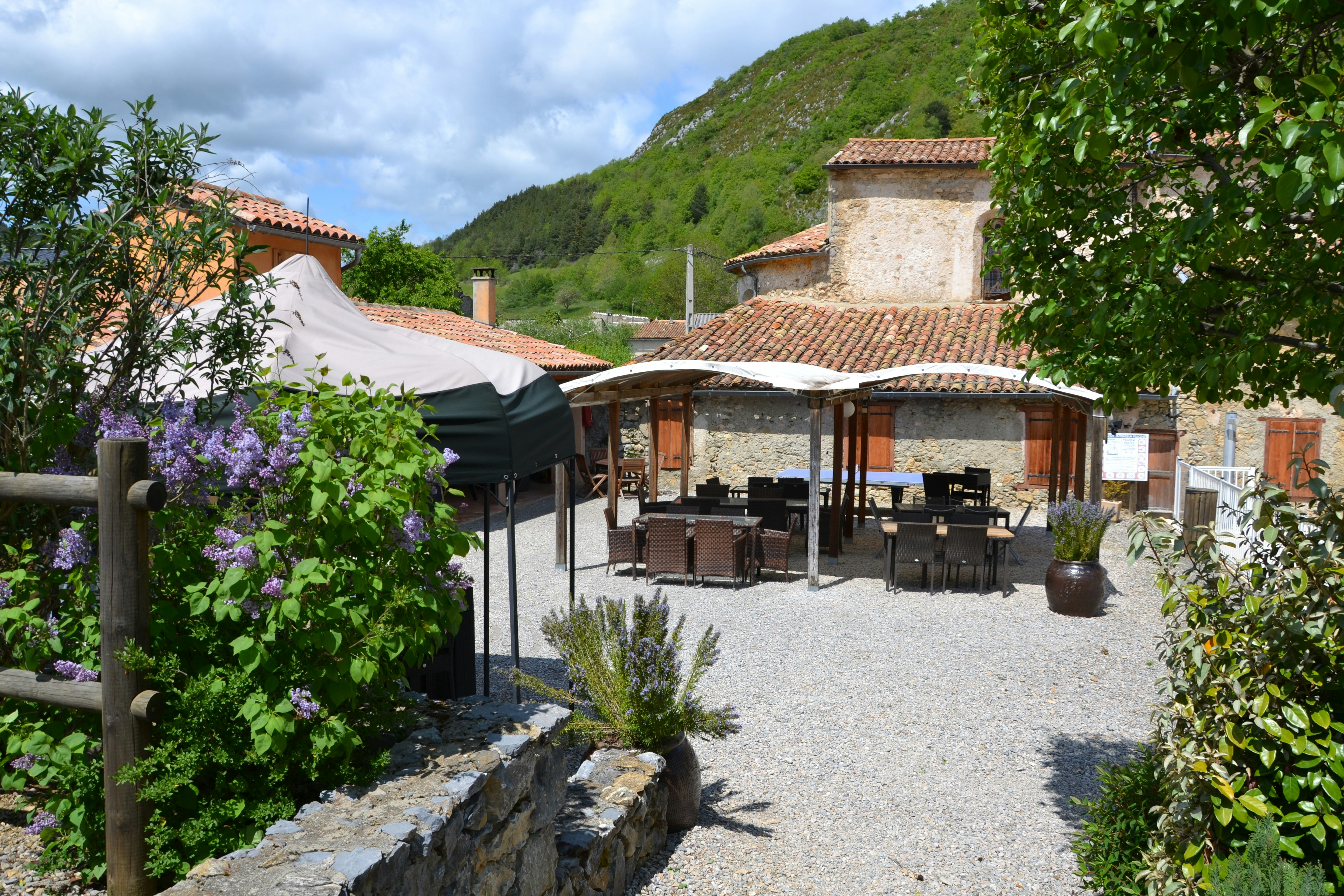 Terrasse mit Holztisch und Stühlen am Campingplatz, Kiesweg, Steinmauer und Gebäude mit Ziegeldach vor grünen Hügeln.