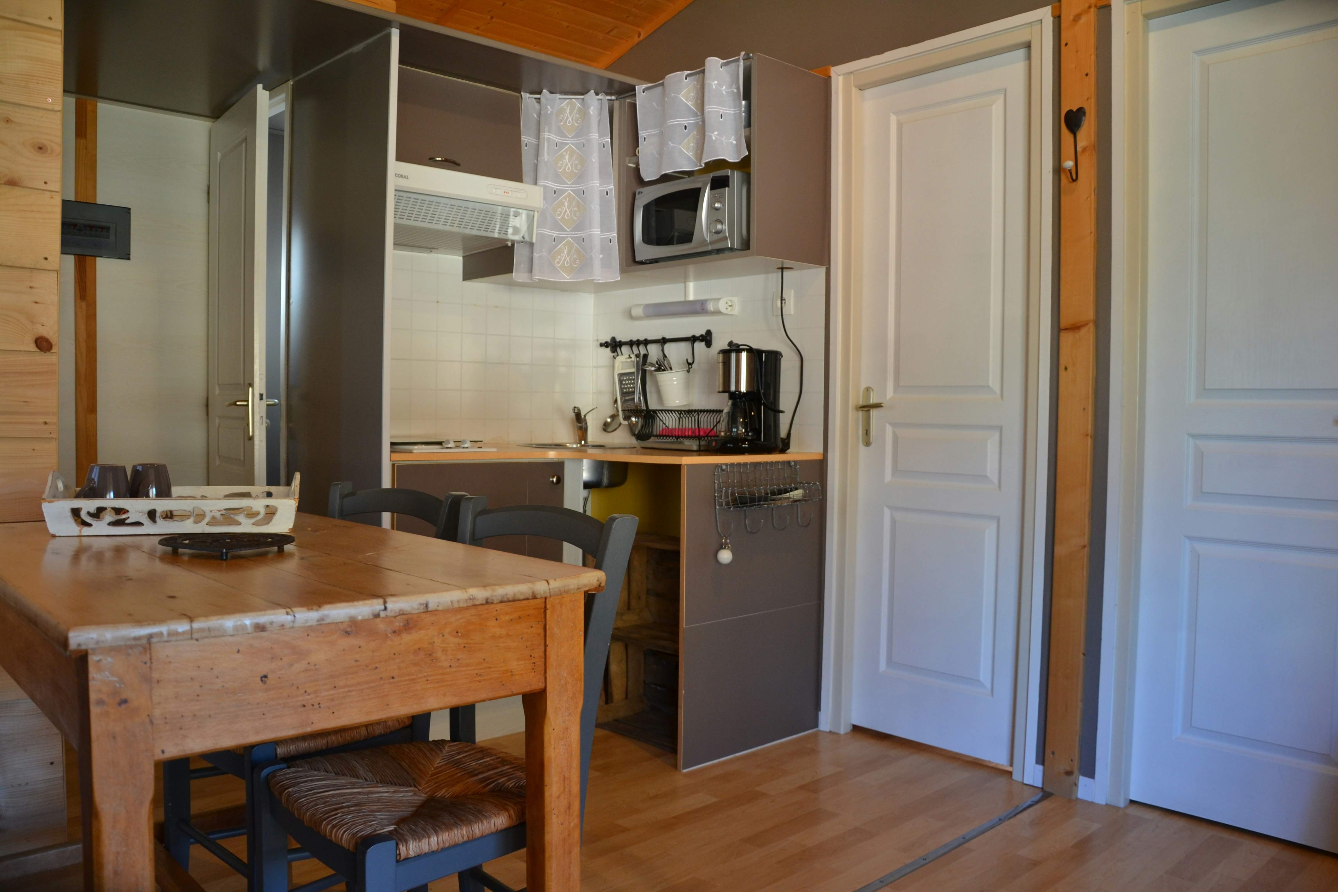 Innenansicht einer Campingküche mit Holztisch, Stühlen, Küchenzeile und Mikrowelle, im Hintergrund offene Tür und Flur.