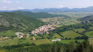 Blick auf ein Dorf im Tal mit Häusern, Wiesen und Wäldern, umgeben von grünen Bergen und Hügeln.