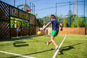Fußballplatz mit Kunstrasen, Tor und Umzäunung; Kinder spielen mit einem gelben Ball vor einem Holzsichtschutz.