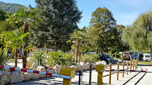 Campingplatzweg mit Steinmauer, Holzgeländer und bunten Sitz-/Liegeelementen, dahinter Bäume und ein geparkter Wagen.
