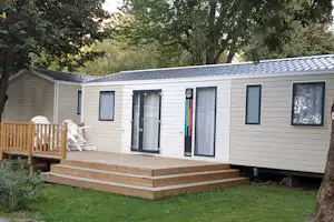 Mobilheim mit Holzterrasse und Sitzbank, seitlich mit Fenstern und Eingangstür, umgeben von Bäumen und Rasen.