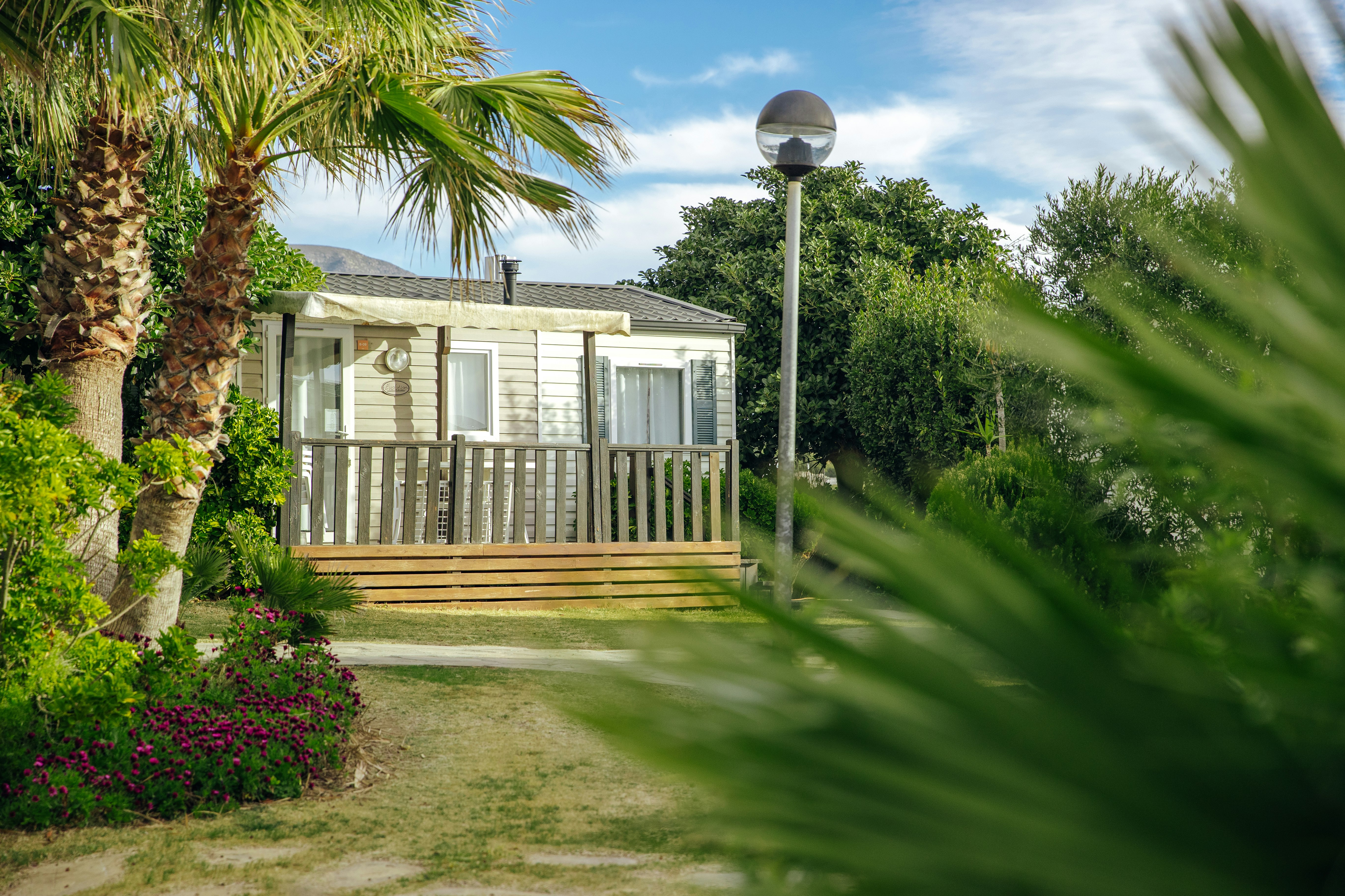 Bungalow mit Holzterrasse, Gartenweg und Laterne, umgeben von Palmen, Sträuchern und bunten Blumenbeeten.