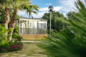 Bungalow mit Holzterrasse, Gartenweg und Laterne, umgeben von Palmen, Sträuchern und bunten Blumenbeeten.