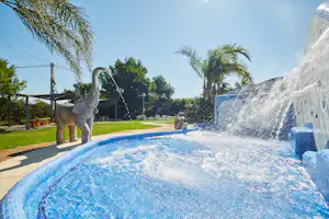 Großer Camping-Pool mit Wasserfontäne und Rutsche, Palmen und Gartenweg im Hintergrund.