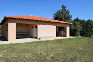 Camping-Bungalow mit Terrasse, offener Veranda und Rasenfläche davor, dahinter Bäume und ein klarer Himmel.