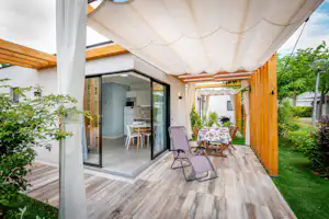 Überdachte Camping-Terrasse mit Sonnenschirm, Liegestuhl, Tisch und Stufen zum Eingang, umgeben von Hecken und Grünpflanzen.