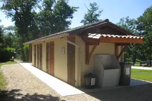 Campinghaus mit Holzüberdachung und Grillbereich, daneben Mülltonnen, gepflasterter Weg und Kiesfläche, umgeben von Bäumen.