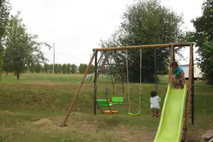 Grüner Rutschenturm mit Schaukeln auf einem Campingplatz, im Hintergrund Wiese und Bäume.