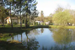 Ruhiger Camping-Teich mit Spiegelung, Bäumen und Büschen am Ufer, im Hintergrund Wohnwagen und Gebäude.