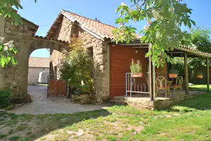 Steinhaus mit Holzveranda, Garten und Sitzgruppe unter einem Vordach, umgeben von Bäumen und gepflastertem Weg.