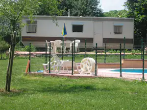 Wohnwagen mit Terrasse und Gartenmöbeln im Grünen, Zaun und Rasen vor einem weißen Gebäude.