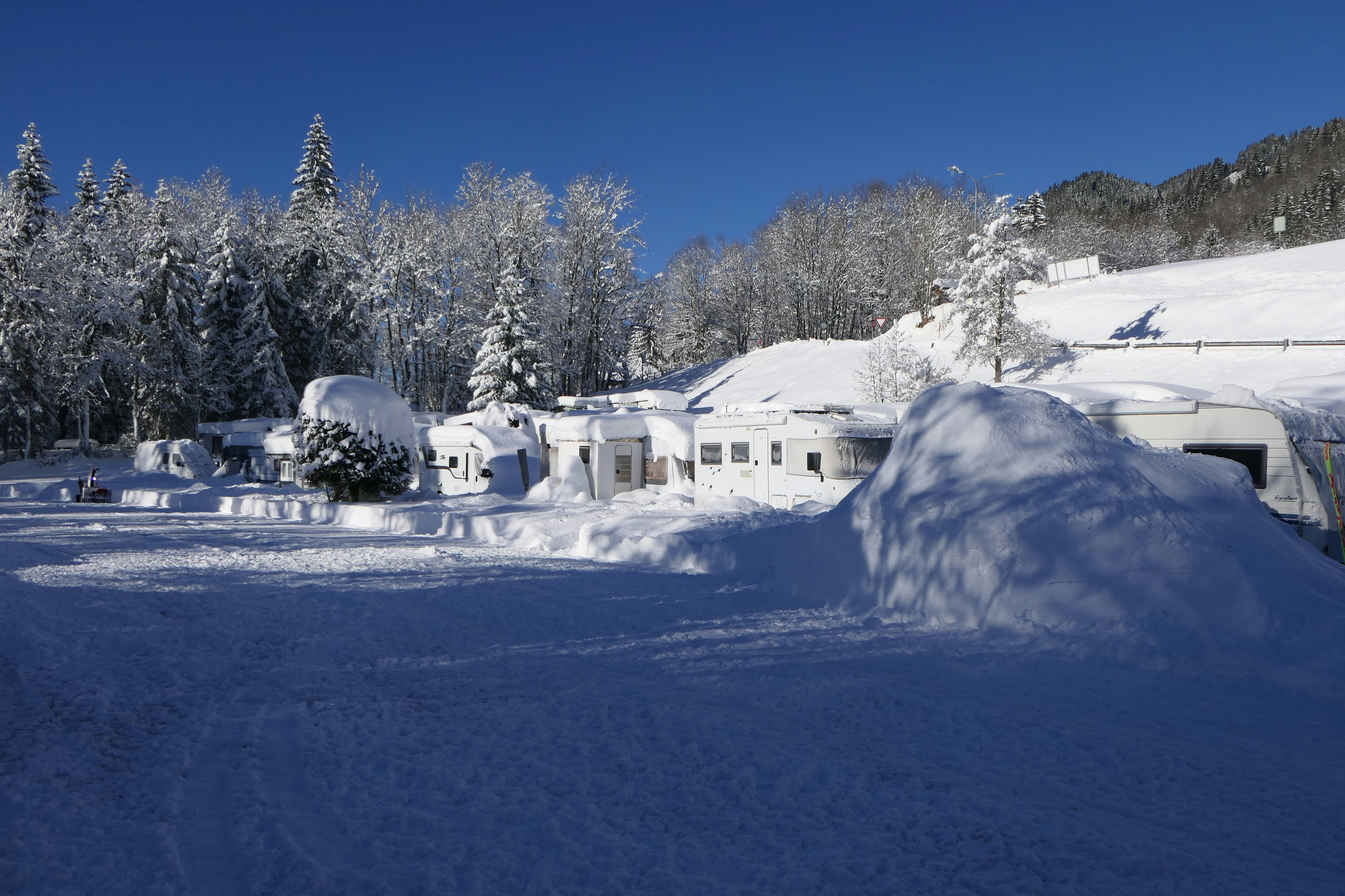 Schneebedecktes Campinggelände mit Wohnmobilen und Zelten, im Hintergrund verschneite Bäume und Berge unter klarem Himmel.