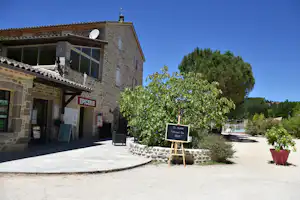 Steinbau mit Ladenbereich „EPICERIE“, Schaukasten und Sitzbank vor Terrasse, umgeben von Bäumen und Gartenbeeten.