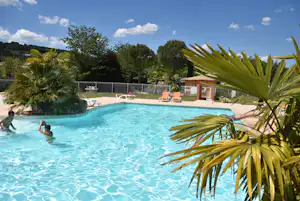 Blauer Campingpool mit Palmen im Vordergrund, Liegen auf der Terrasse und Zaun im Hintergrund, umgeben von Bäumen.