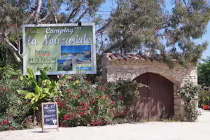 Werbeschild „Camping La Nouzarede“ am Eingang, mit Blumenbeeten, Bäumen und einem großen Steingebäude mit Holztor.