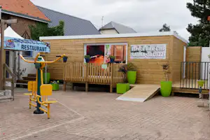 Holzterrasse am Campingplatz mit Schild „Restaurant“, Bereich „Espace Bien-Être Sauna-Jacuzzi“ und Liegestuhl, daneben Fitnessgerät.