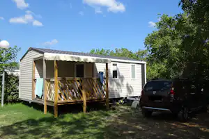 Wohnwagen mit überdachter Veranda und Holzgeländer, daneben ein Auto auf dem Campingplatz, Rasen und Bäume im Hintergrund.