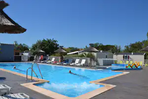 Schwimmbecken mit Liegen am Campingplatz, im Hintergrund kleine Gebäude und Sonnenschirme, daneben ein Spielbereich mit bunten Reifen.