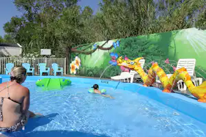 Camping-Pool mit Wasserspielzeug, grünem Poolrand und bunten Aufblasfiguren an der Wand, daneben ein Liegestuhl.