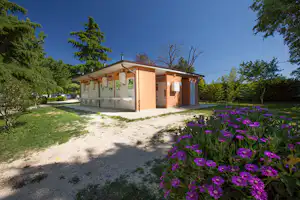 Sanitärgebäude auf dem Campingplatz mit Waschbereich, Schiebetür und Blumenbeet im Vordergrund, umgeben von Bäumen und Rasen.