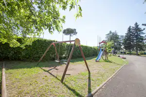 Spielplatz auf dem Campingplatz: Schaukelgestell und blaue Rutsche neben Hecken, Rasenfläche und Weg.