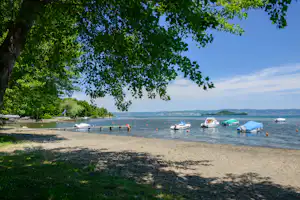 Blick auf den See mit Booten und Stegen, Sandstrand und grünen Bäumen im Vordergrund.