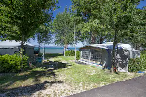 Zelt und Wohnwagen auf einem Campingplatz am See, umgeben von Bäumen und Grasfläche.