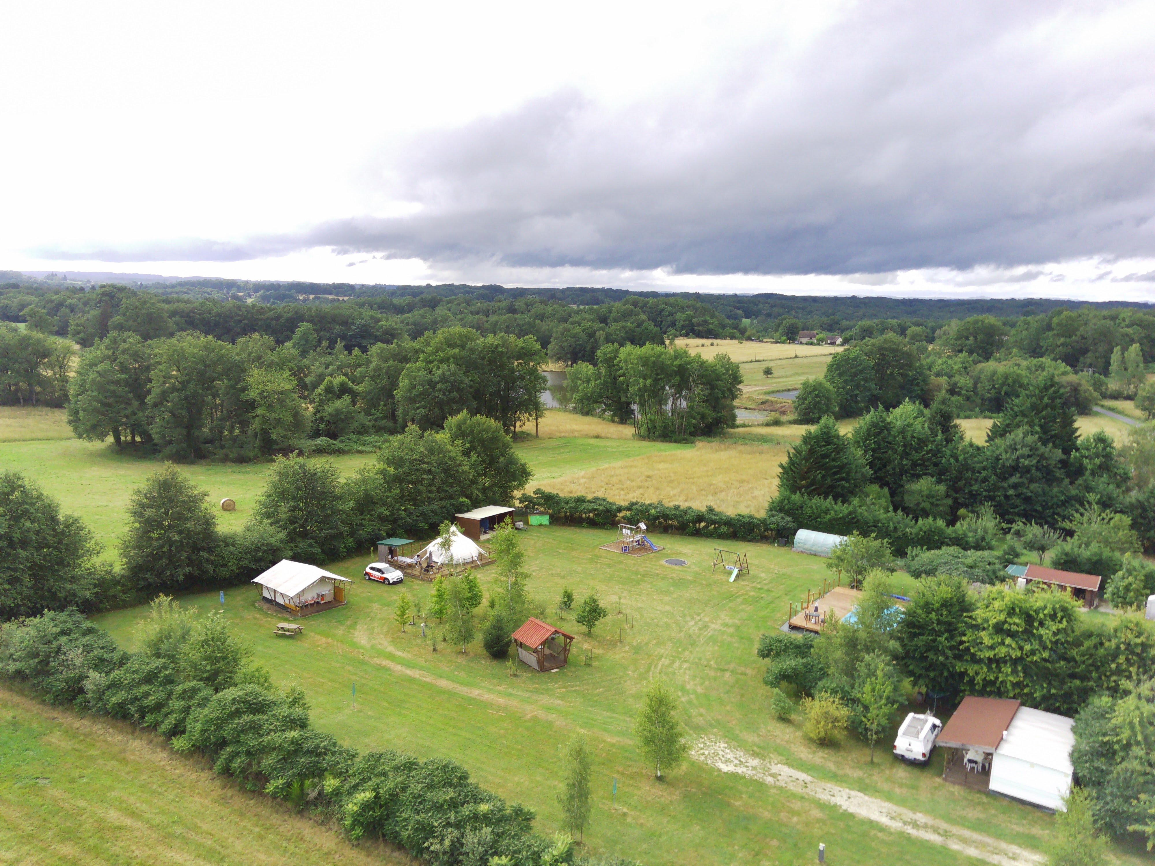 Campingplatz auf einer Wiese mit Zelten und Wohnwagen, umgeben von Bäumen und grünen Feldern, Blick von oben.