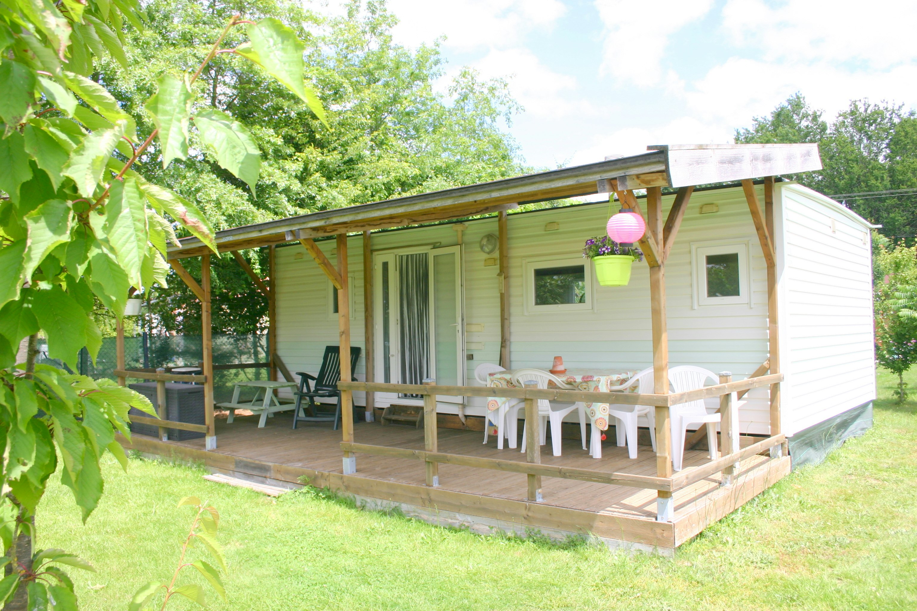 Holzbungalow mit Veranda, Tisch und Stühlen, umgeben von Rasen und Bäumen im Campinggarten.