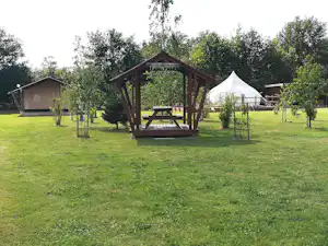 Spielplatz mit Holzunterstand und Tisch, daneben ein weißes Zelt auf einer grünen Wiese, im Hintergrund Campinghäuser und Bäume.
