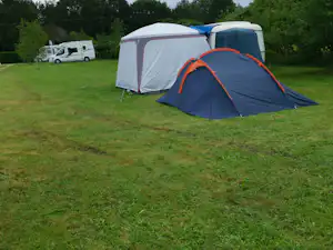 Campingzelt auf einer grünen Wiese, daneben ein Wohnmobil und Bäume im Hintergrund.