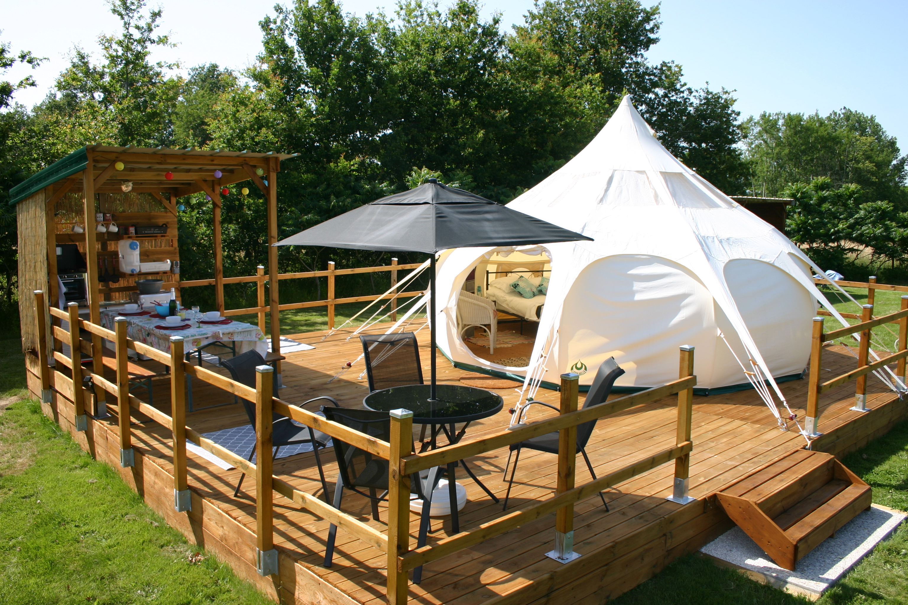 Weiße Zelte auf einem Holzpodest in einem Campingbereich, mit Tisch, Stühlen und Holzumzäunung, umgeben von Bäumen.
