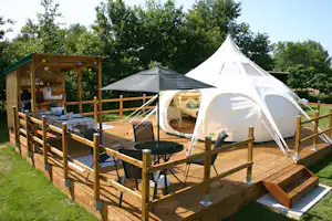 Weiße Zelte auf einem Holzpodest in einem Campingbereich, mit Tisch, Stühlen und Holzumzäunung, umgeben von Bäumen.